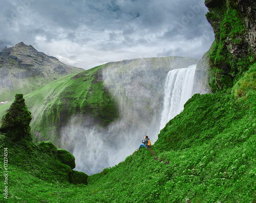 Girl in waterproof clothing sits on the cliff on background of Skogafoss waterfall in Iceland. View from above photo