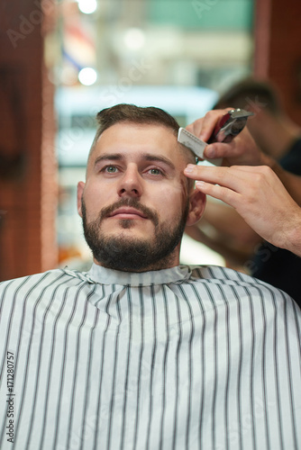 Vertical shot of a handsome young bearded man sitting relaxed in a chair at the barbershop while getting his hair styled. 