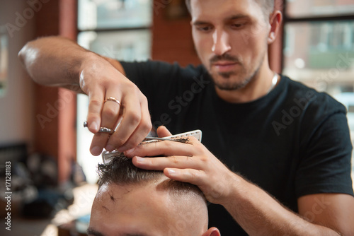 Selective focus on the hands of a professional barber with scissors and comb styling hair of his client. 