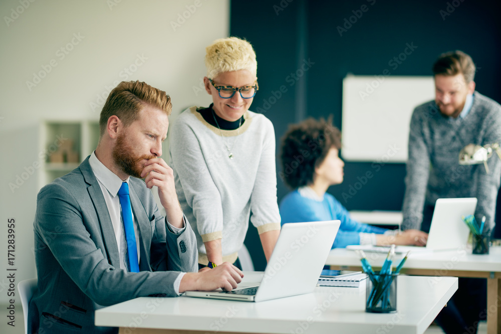 Obraz premium Businessman sitting and using laptop in modern office while his colleague standing and helping him to solve a problem