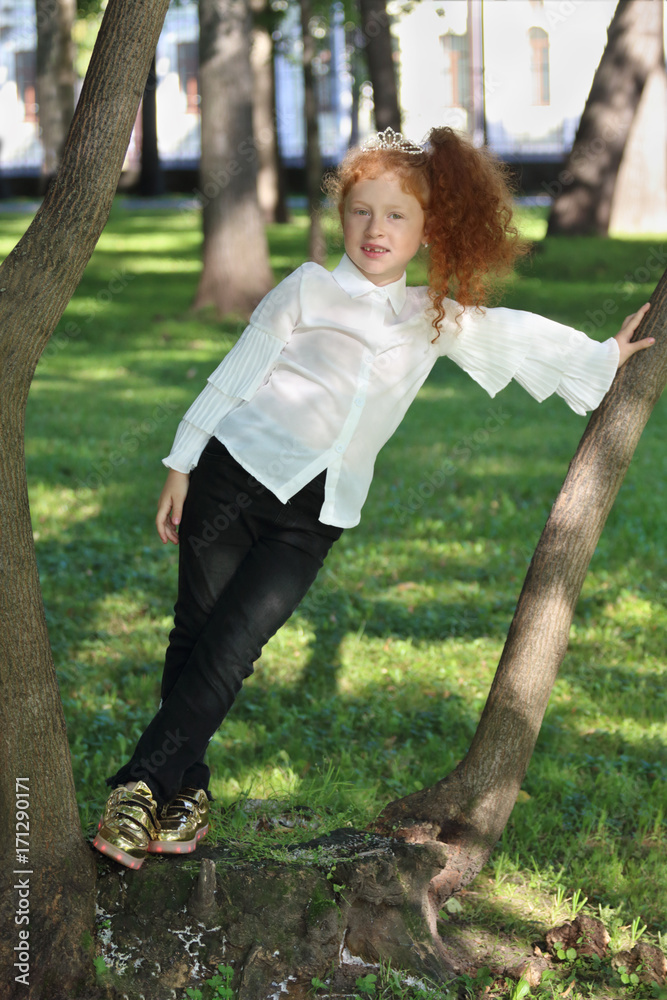 Φωτογραφία Little girl with red hair in jeans poses near tree in summer green park