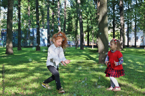 Two little girls play with soap bubbles near trees in summer green park
