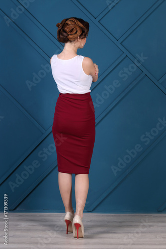 Beautiful woman with hairdo and in skirt poses in blue studio, back view