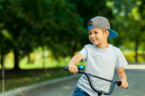 Little boy learns to ride a bike in the park near the home. Portrait of a cute kid on bicycle. Happy smiling child in cap riding a cycling.