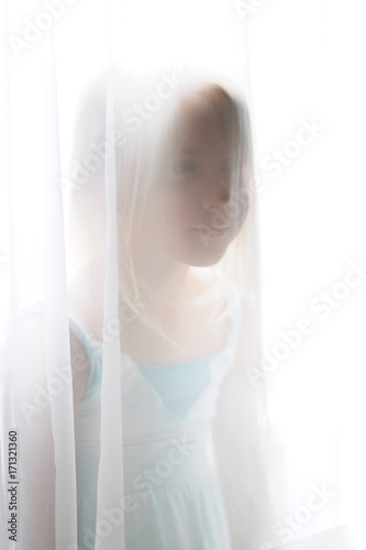 A nice little blond girl stands by the window behind a transparent curtain