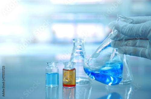 glass flask vial with blue and orange  liquid in chemical science laboratory with scientist hand with glove background