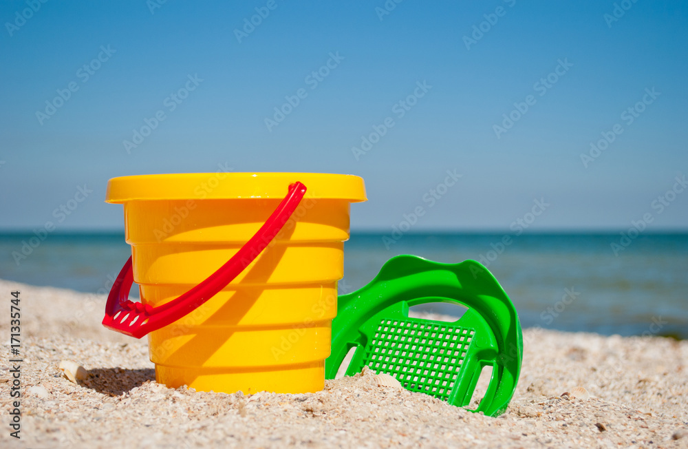 Yellow childrens bucket with red handle and plastic green sieve left on a blue sea and blue sky background yellow sand beach sea shore seashells summer vacation sunny summer day, bright sun, baby toys Yellow childrens bucket with red handle and plastic green sieve left on a blue sea and blue sky background yellow sand beach sea shore seashells summer vacation sunny summer day, bright sun, baby toys