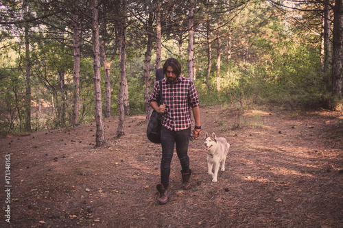 Handsome bearded man with guitar bag and husky dog walking in the woods at summer times 