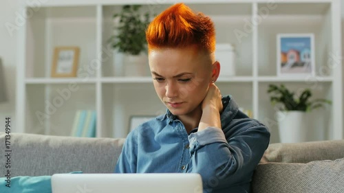 Portrait of red hair woman using laptop for chatting while feeling anoying neck ache at home in the living room. photo