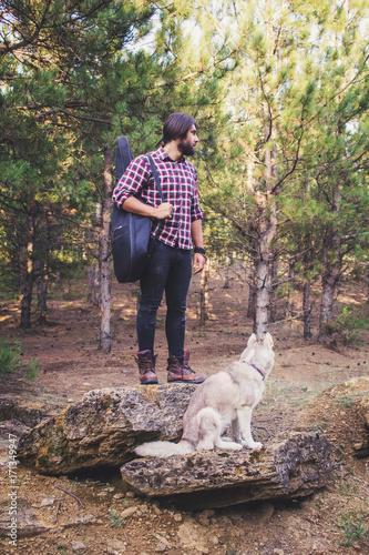 Handsome bearded man with guitar bag and husky dog walking in the woods at summer times  