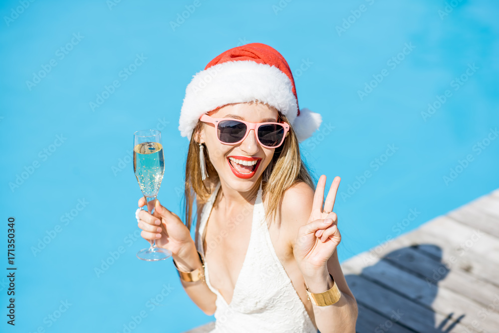 Obraz premium Young woman in Christmas hat and swimsuit celebrating New Year with glass of wine at the swimming pool. Winter vacation concept