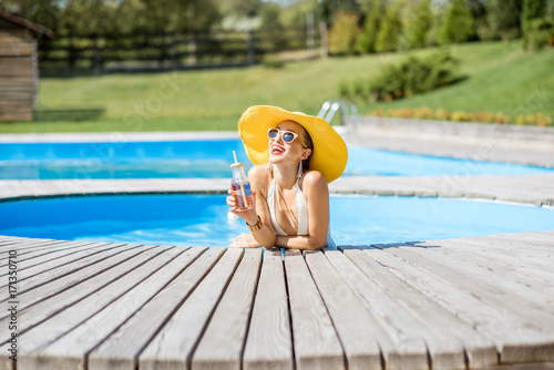 Young woman in swimsuit with big yellow sunhat relaxing with a bottle of fresh drink sitting on the poolside outdoors