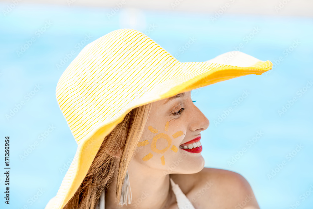 Close-up portrait of a young woman in yellow hat with sun painting on her cheek on the blue water background