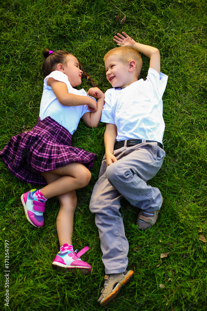 Fototapeta premium Enjoying summer time. Top view of two cute little children holding hands behind head and looking at each other with smile while lying on the green grass together