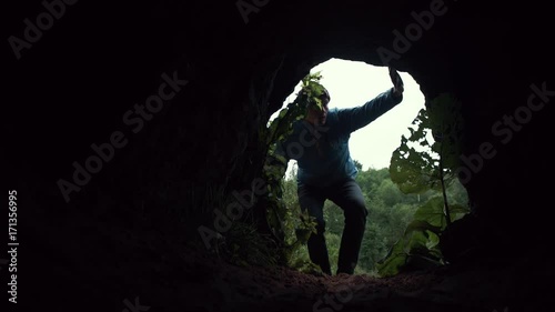 Young hiker crawls on all fours in darkness to exits from cave and stands up. Traveler inside the cave. A look from the cave. Deep forest at background.