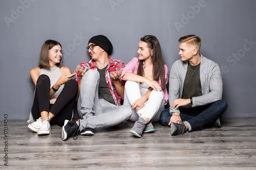 Group of beautiful students in casual clothes using gadgets, talking and smiling while sitting together on the floor