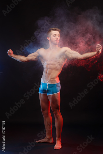 Young handsome muscular man bodybuilder with perfect abs, shoulders,biceps and triceps demonstrating huge muscles in the smoke, full length portrait © Vadym