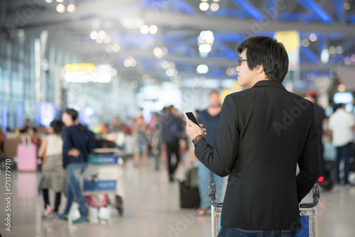 Young asian man with his suitcase luggage using smartphone while waiting for airline flight in the international airport terminal, business travel and online check in concepts