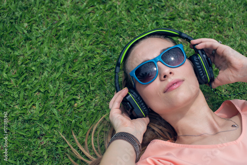 Pretty girl with headphones and sunglasses is lying on the grass in the park (sunny day) © Alina