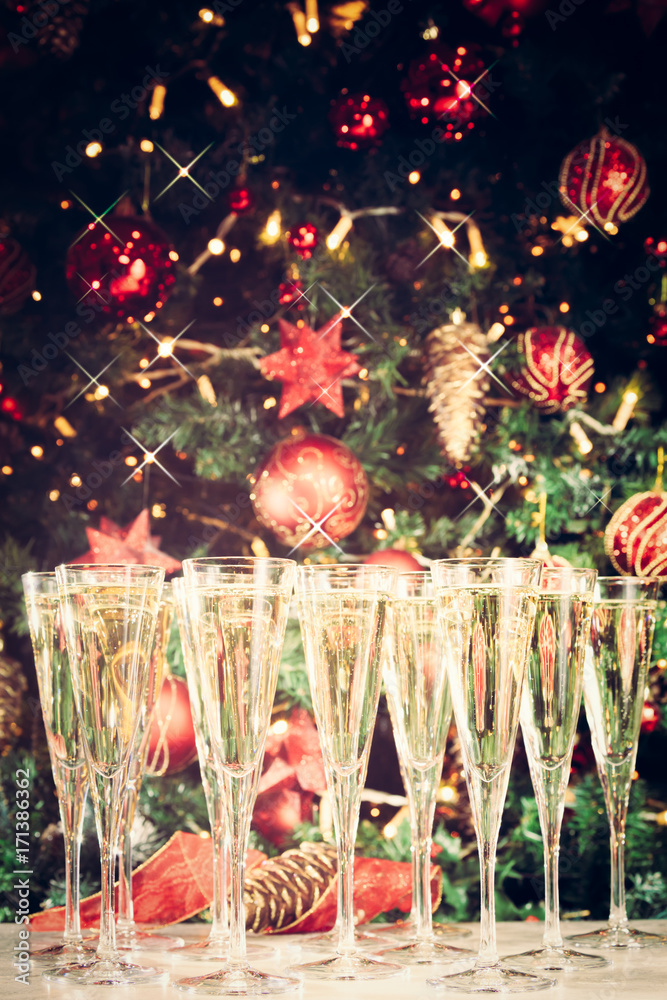 Fototapeta premium Glasses of champagne with Christmas tree background and sparkles. Many glasses for party. Holiday season background. Traditional red and green Christmas decoration