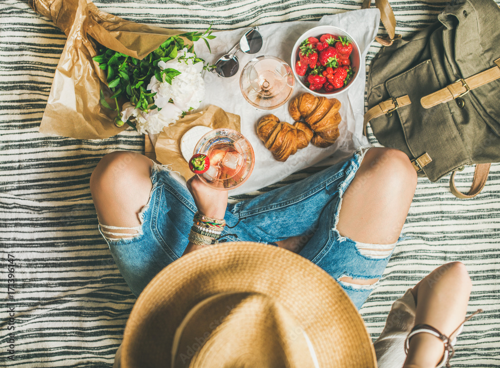 French style romantic picnic setting. Woman in shabby jeans and hat with  glass of ice rose wine, strawberries, croissants, brie cheese, sunglasses,  peony flowers, top view. Outdoor gathering concept foto de Stock