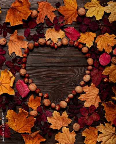 Flat lay frame in the form of a heart from autumn crimson and yellow leaves, hazelnuts and walnuts on a dark wooden background.