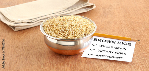 Brown rice, which is a whole grain and healthy food, in a copper bowl, with a note of some of its benefits, on a wooden background.