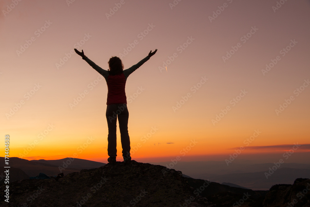 Sunset Silhouette of attractive woman staying and looking happy on top of the mountain. Freedom, adventure and leisure vacation concept.