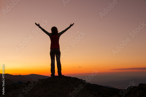 Sunset Silhouette of attractive woman staying and looking happy on top of the mountain. Freedom, adventure and leisure vacation concept.
