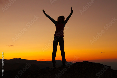 Sunset Silhouette of attractive woman staying and looking happy on top of the mountain. Freedom, adventure and leisure vacation concept.
