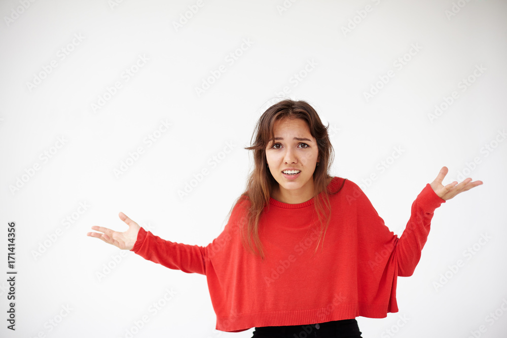 Pretty female in red sweater posing at studio wall shrugging shoulders with open palms, feeling clueless or confused, her look and gesture expressing indifference as if she saying: Who cares? So what?