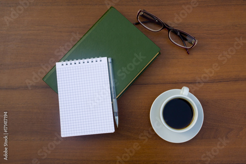 Cup of coffee, book, notepad, pen and eyeglasses on wooden desk