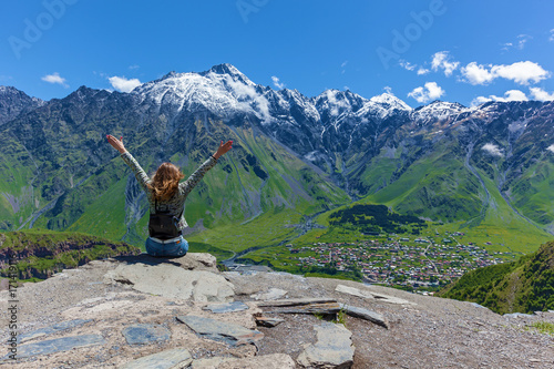 girl sits on cliff's edge and looks at the tops of mountains near the village of Gergeti in Georgia, under Mount Kazbegi. Girl looks at the tops of mountains