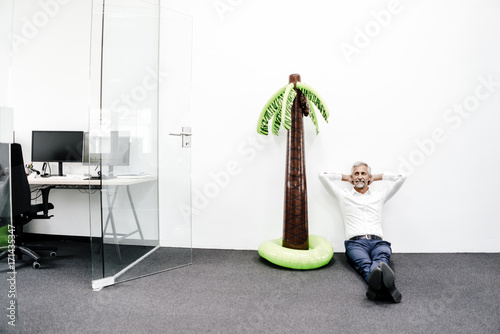 Smiling mature businessman sitting next to inflatable palm tree in office