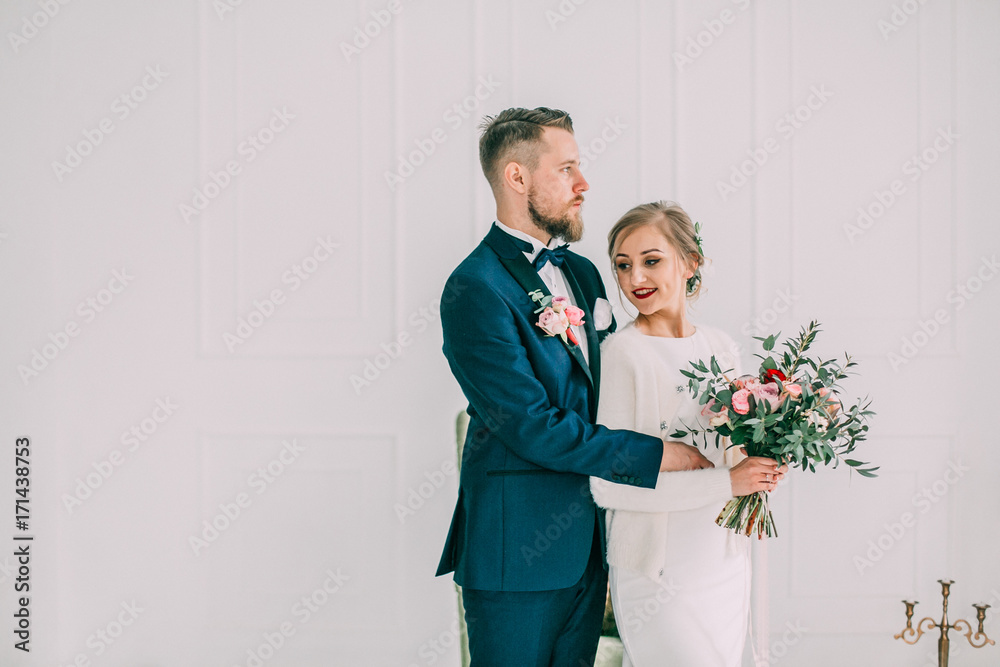 Bride in beautiful dress and groom in black suit sitting on sofa indoors in white studio interior like at home. Trendy wedding style .bride hugging Groom