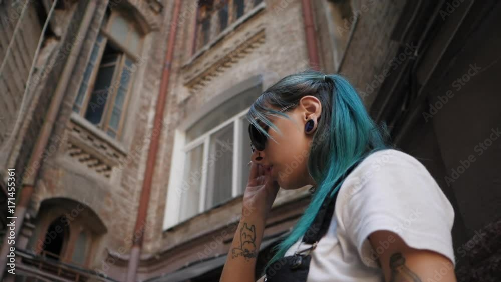 SLOW MOTION: Homeless emo girl smoking cigarette.Street punk or hipster in sunglasses with blue dyed hair,piercing,lenses,tattooed,ears tunnels and unusual hairstyle stands in backyard. SLOW MOTION: Homeless emo girl smoking cigarette.Street punk or hipster in sunglasses with blue dyed hair,piercing,lenses,tattooed,ears tunnels and unusual hairstyle stands in backyard.
