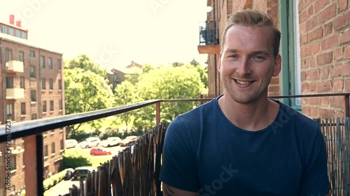 Smiling casual man wearing a blue tshirt sitting down on his balcony outdoors on a sunny morning.