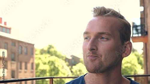 Smiling casual man wearing a blue tshirt sitting down on his balcony outdoors on a sunny morning.