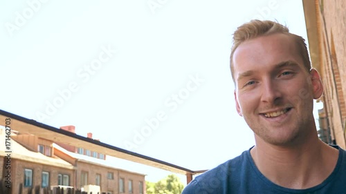 Smiling casual man wearing a blue tshirt sitting down on his balcony outdoors on a sunny morning.