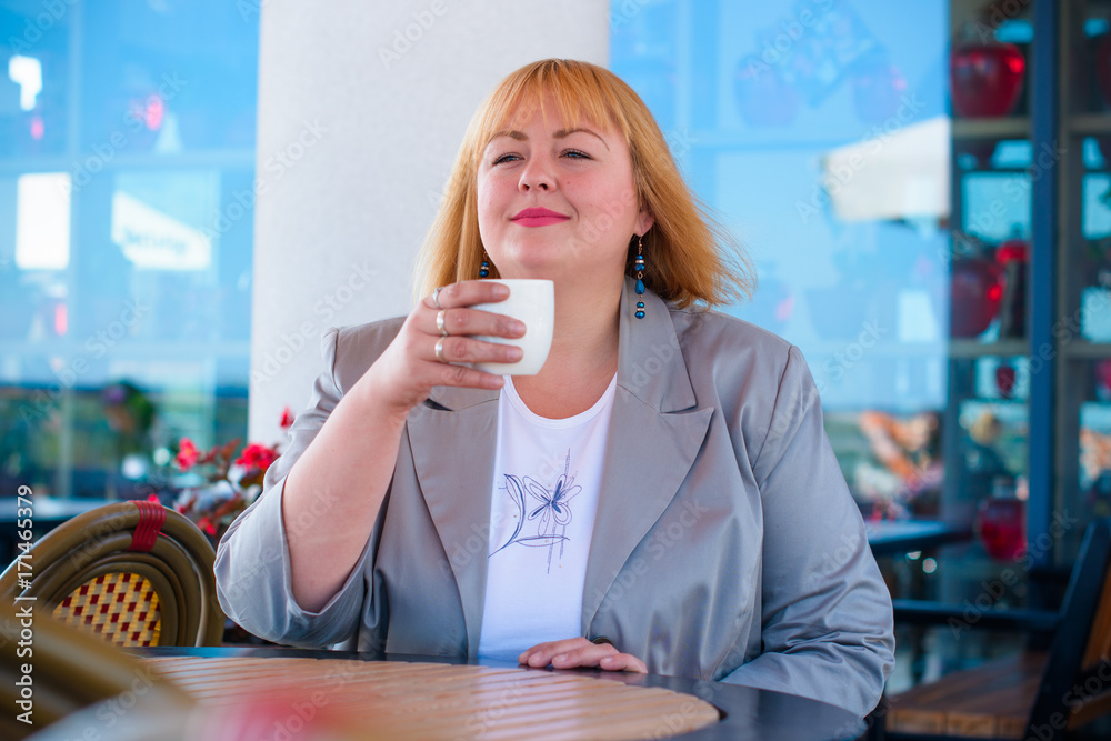 Cheerful plump lady is sitting at the table in cafe and drinking tea. Young pretty funny woman having lunch at a cafe on the street