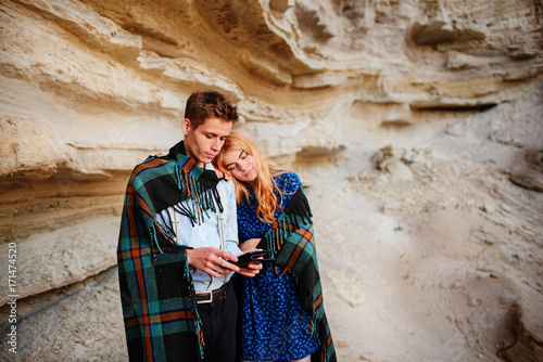 Beautiful woman and handsome man wrapped in a blanket. They are smiling and looking at the screen of a tablet on the background of a sand quarry