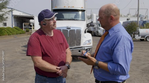 Truck driver talks to official, insurance agent or delivery manager, in shirt and tie who asks him to sign an electronic notepad. Medium shot in front of parked white truck on summer day. 4K
