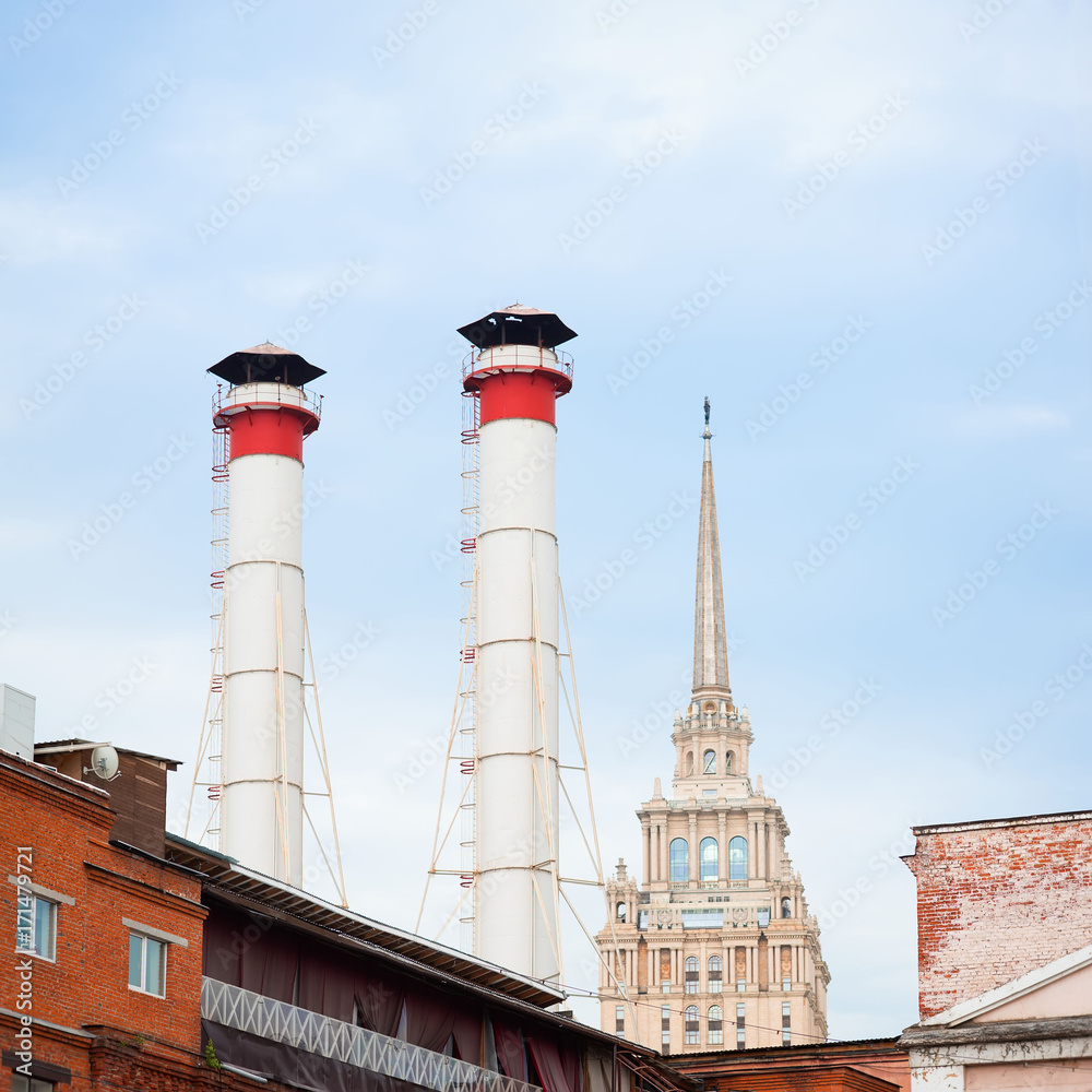 Mix of two pipes and buildings of different times against blue sky. Old Industrial building of the factory from red brick, smoking chimneys, tower residential building