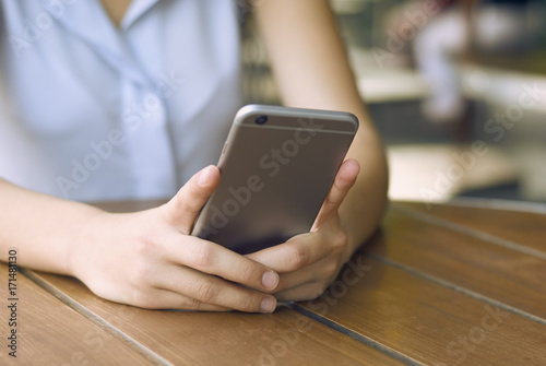 the girl is holding a dark-colored phone in daylight against a light brown wooden table. background blur