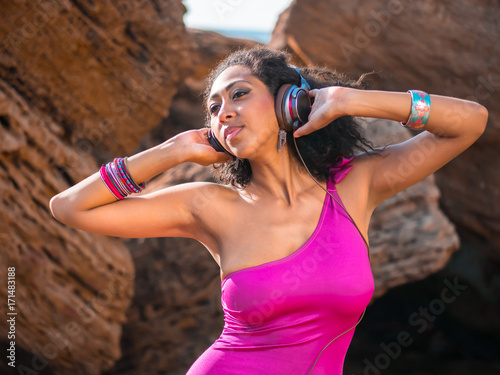 Young pretty black afro female wearing pink swimsuit and brown headphones sitting on tropical rocks beach near sea,enjoying beautiful summer day.Carefree, music concept.