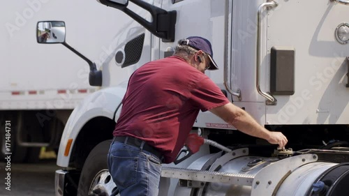 Truck driver walks over with hose and starts filling tank up with diesel fuel with other trucks in background. Exterior medium shot, summer sunlight, real time 4K