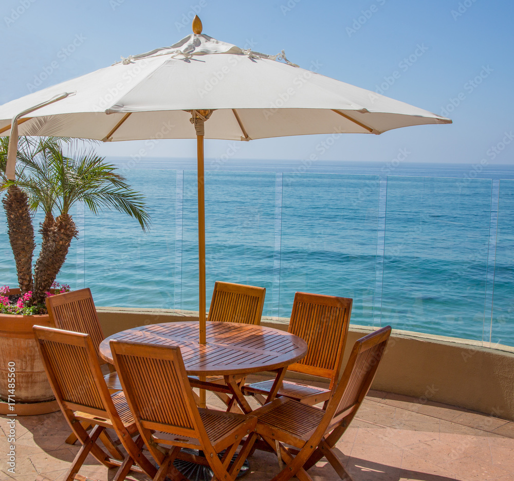 Oceanfront Patio / Wood table, chairs and umbrella on a large patio overlooking the blue ocean waters.