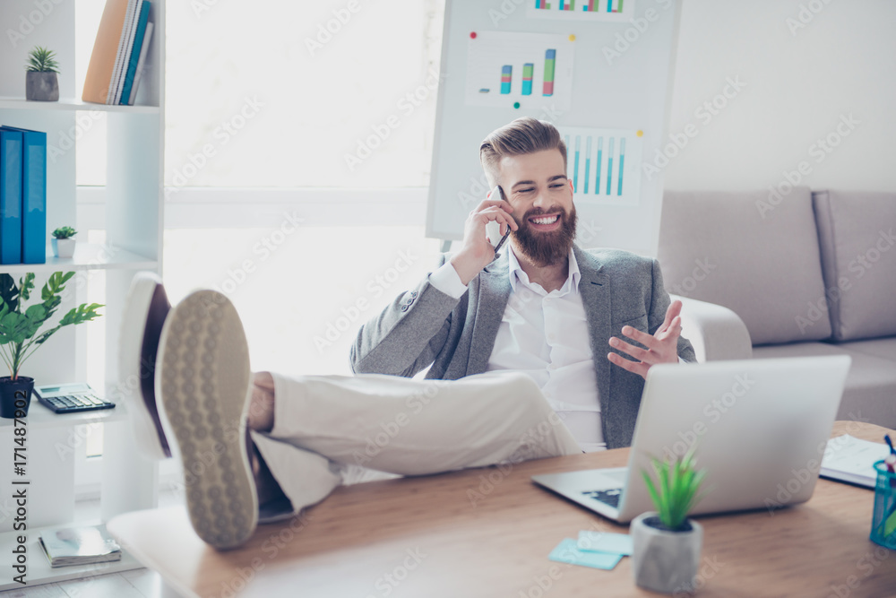 Handsome young guy in formalwear talking on mobile phone and telling about his achievement, sitting at the office with feet on the desk top, gesturing