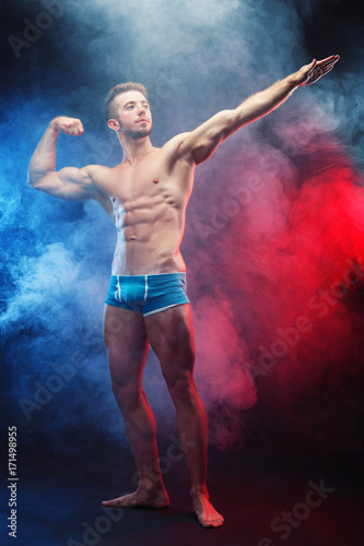 Young handsome ripped muscular man bodybuilder with perfect abs, shoulders,biceps and triceps demonstrating huge muscles in the smoke, full length portrait