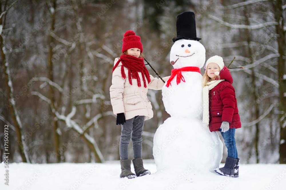 Two adorable little girls building a snowman together in beautiful winter park. Cute sisters playing in a snow. Two adorable little girls building a snowman together in beautiful winter park. Cute sisters playing in a snow.